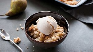 Pear crisp with oats in a black bowl topped with vanilla ice cream on a black table beside a spoon and a pear