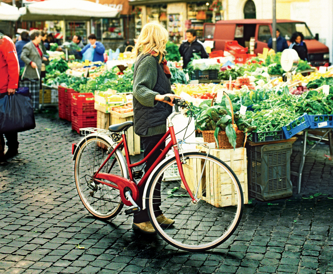 A woman standing with a red bicycle looking at vegetables in farmers' market