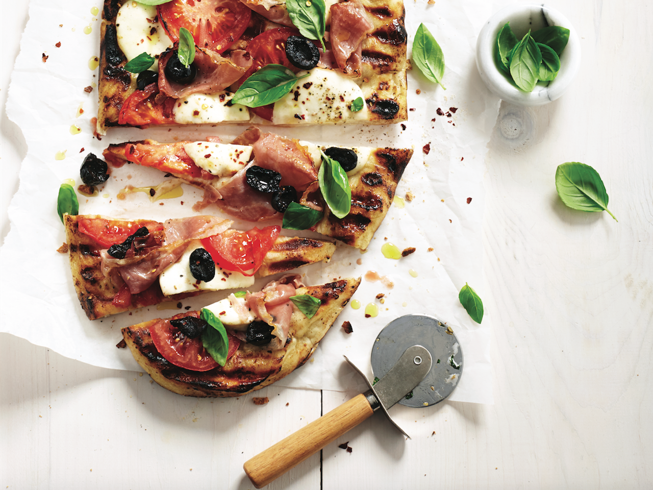 Grilled pizza recipe: Partly sliced grilled pizza on a wooden cutting board, topped with cheese, basil and prosciutto.