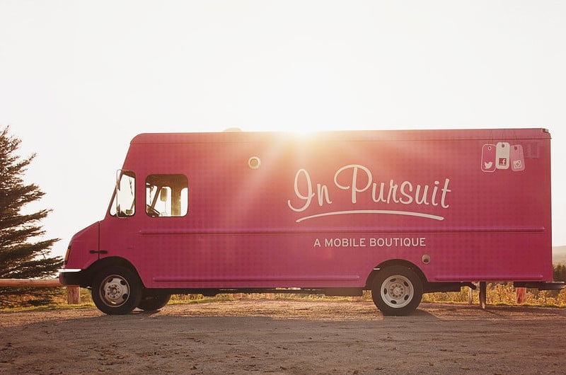 A pink truck sits on a sandy road as the sunset. It says 'In Pursuit - A Mobile Boutique' on the side.
