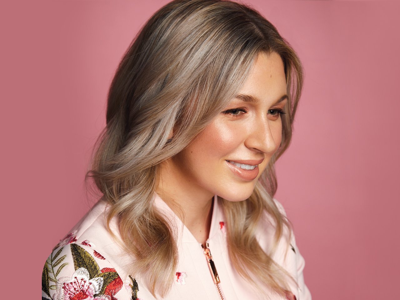 A woman with blonde, voluminous curls on a pink background to illustrate an article on volumizing hair tips for fine hair. 