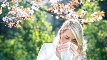 Blonde woman sneezing into a tissue outdoors.