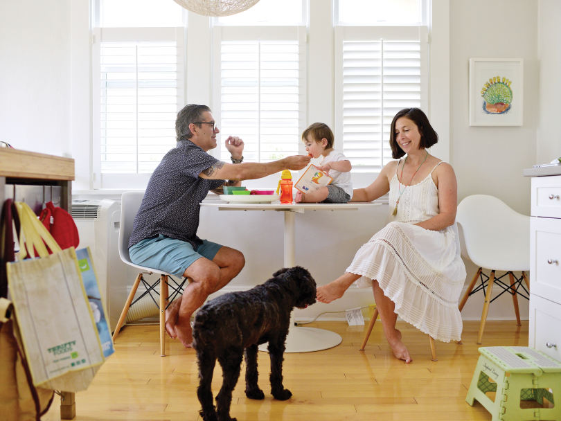 A family sits at the dining table, the child is sitting on the table as a dog licks the mom's toes.
