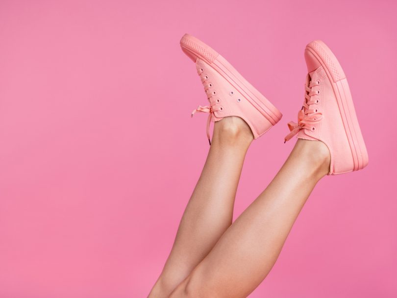 Cropped close-up image view photo of slim shaven legs wearing pink converse-style sneakers isolated over pink pastel background