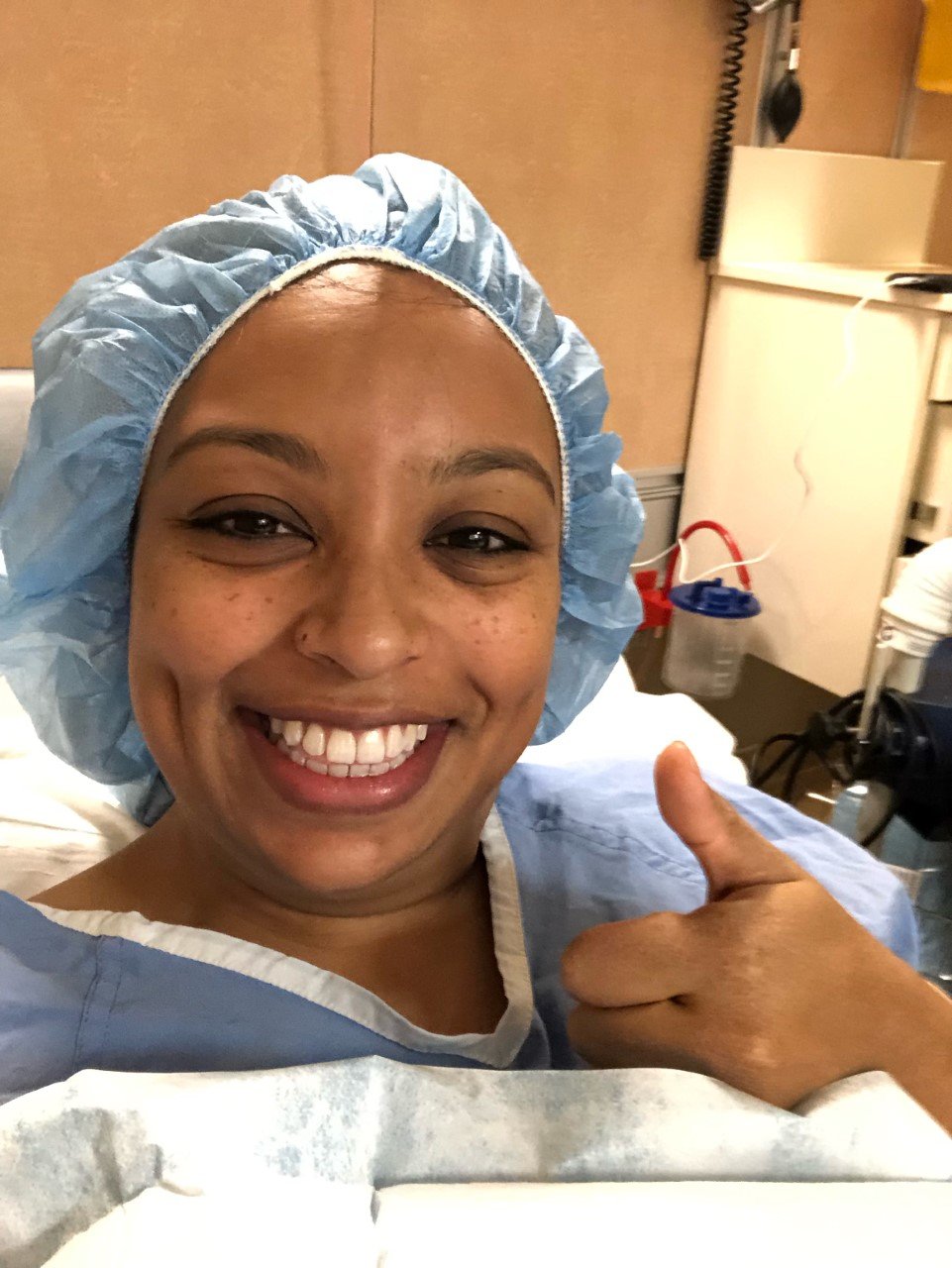 a woman wearing pre-surgery scrubs and hair net gives the camera thumbs up