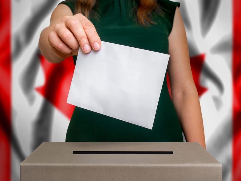 The hand of woman putting her vote in the ballot box. Flag of Canada on background.