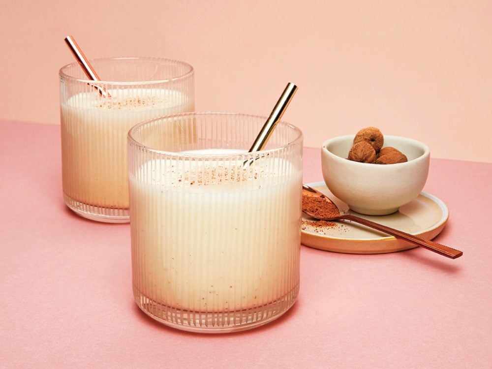 two tumblers of vegan eggnog garnished with cinnamon sticks sitting on a light pink background
