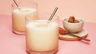 two tumblers of vegan eggnog garnished with cinnamon sticks sitting on a light pink background