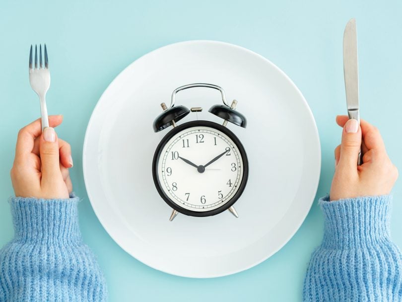 Intermittent Fasting feature image shows an overhead shot of an alarm clock resting face-up on a white plate with hands holding cutlery on either side of the plate