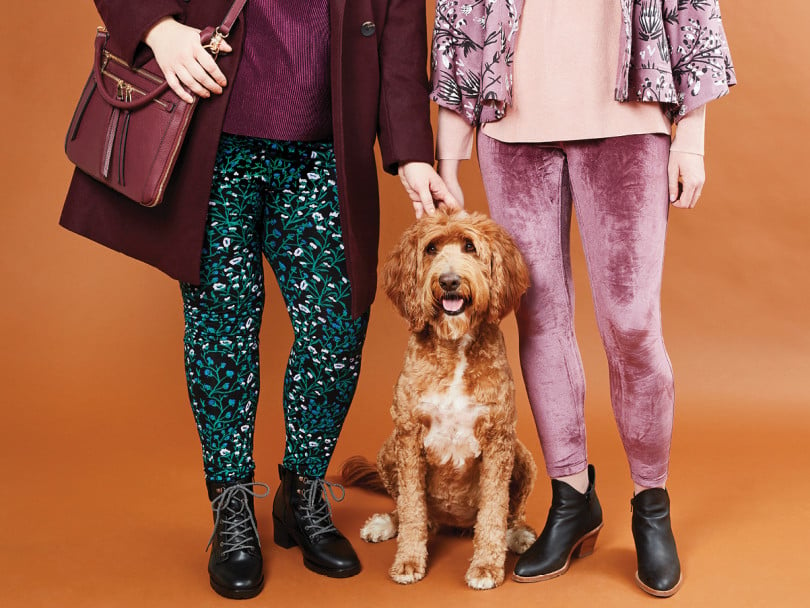 lower half of two women standing against an orange background, one wearing dark floral-print leggings and another wearing pink velvet leggings