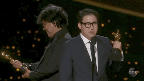 GIF of Bong Joon Ho and Han Jin Won accepting the Oscar for Best Screenplay, Bong is smiling gleefully at the award while Han speaks at the mic