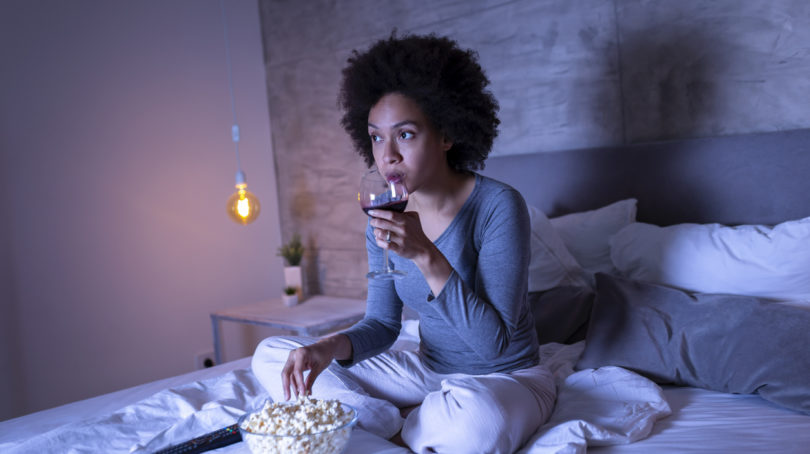 woman sitting on bed, drinking wine, eating popcorn and watching television