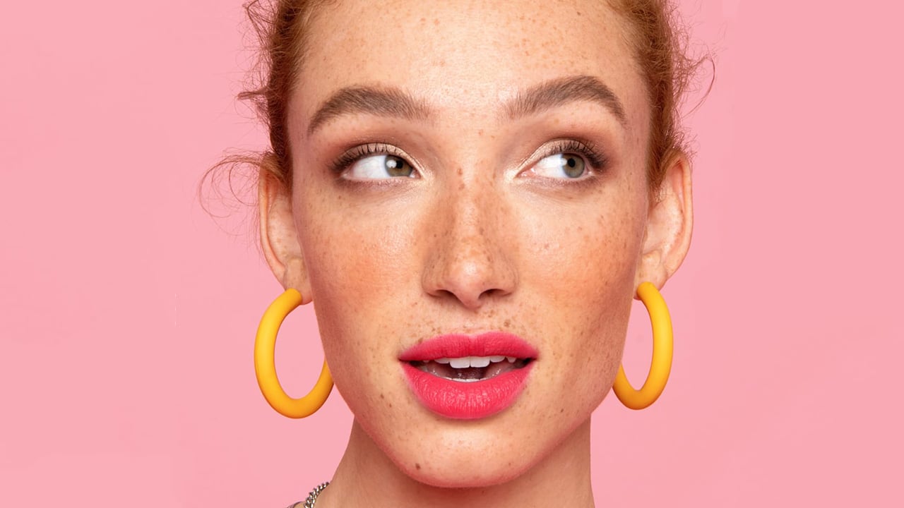 A woman wearing bright pink lipstick and yellow earrings against a pink background for a post on eyebrow makeup products.