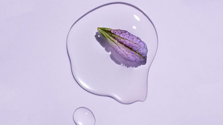 A drop of bakuchiol-infused serum from skincare brand Herbivore against a purple background, with a purple flower petal floating in the drop.