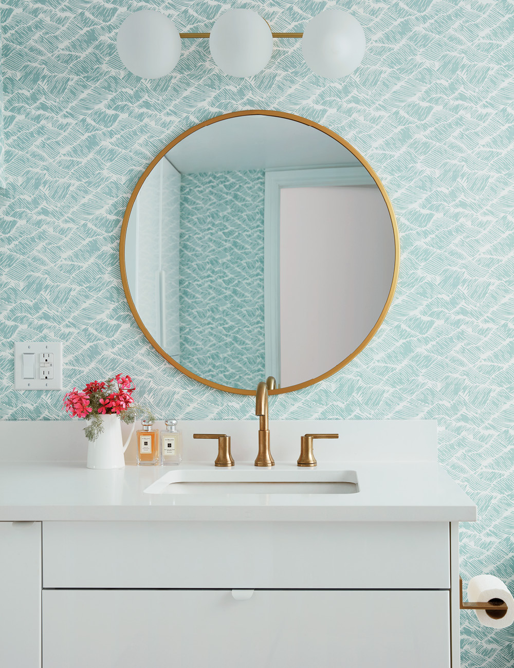 A white bathroom vanity with gold hardware accents and a round mirror on turquoise-patterned wallpaper.