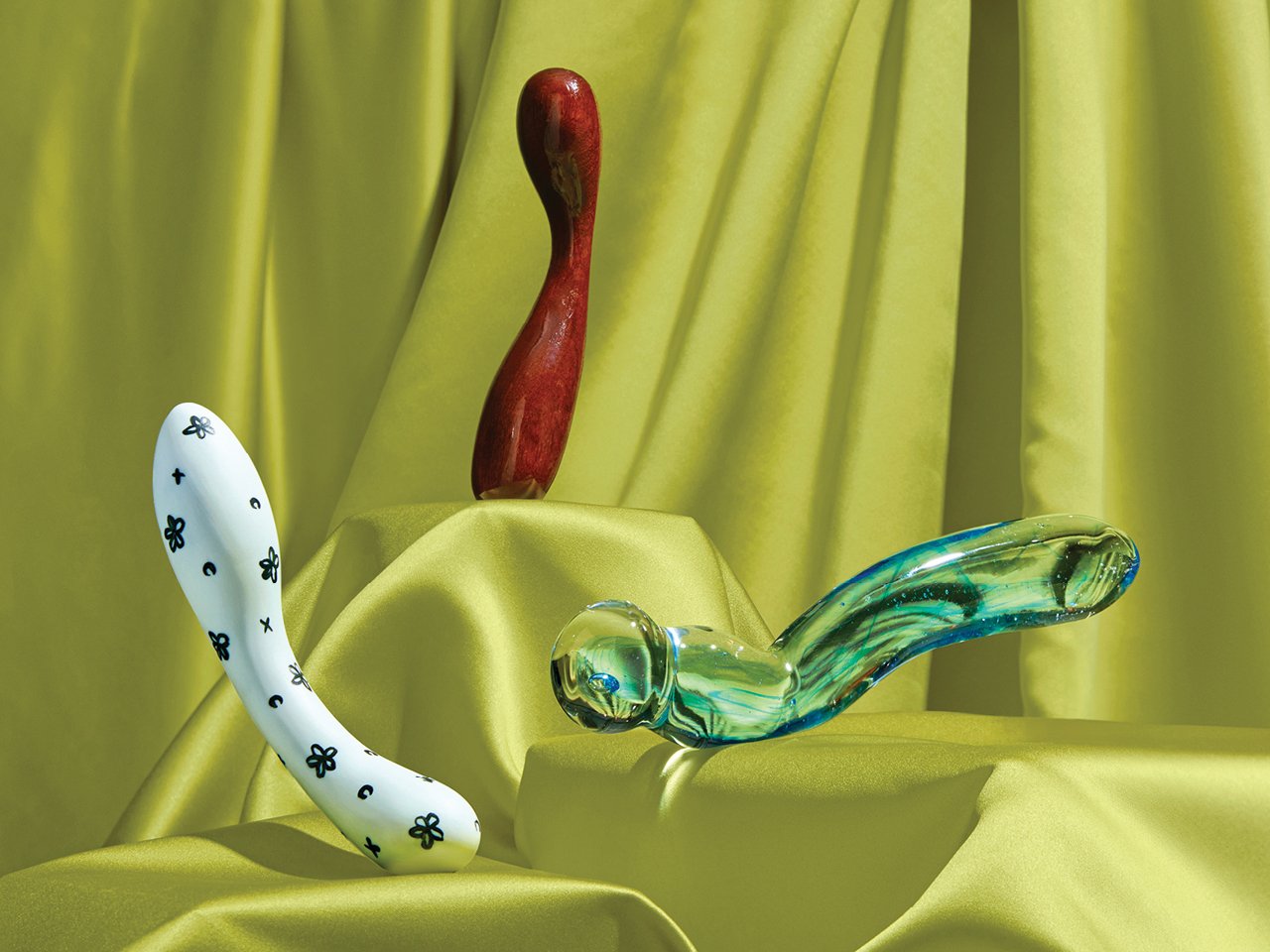 Three dildoes—one white with black flowers, one cherry wood and one green glass—against a chartreuse curtain backdrop