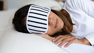 A woman wearing a striped sleep mask lying on her side on a pillow for a piece on the best pillows for sore necks and neck pain, the best pillows for neck pain