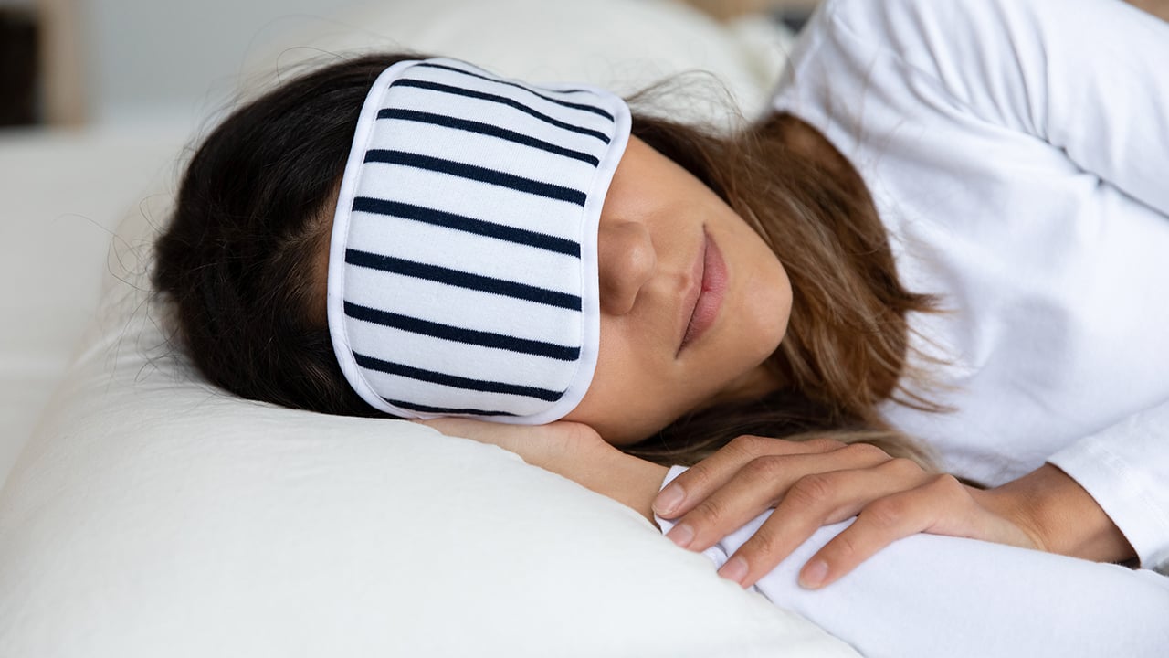 A woman wearing a striped sleep mask lying on her side on a pillow for a piece on the best pillows for sore necks and neck pain