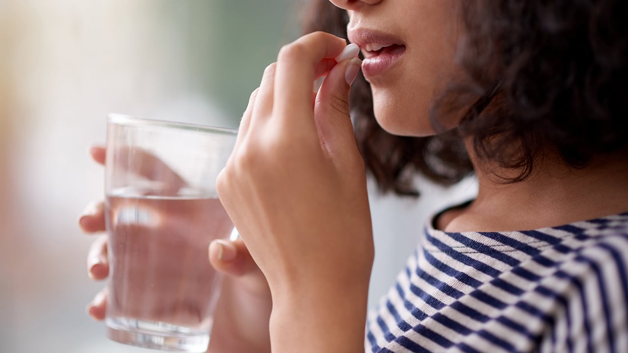 A cropped photo of a woman taking a pill with a glass of water.