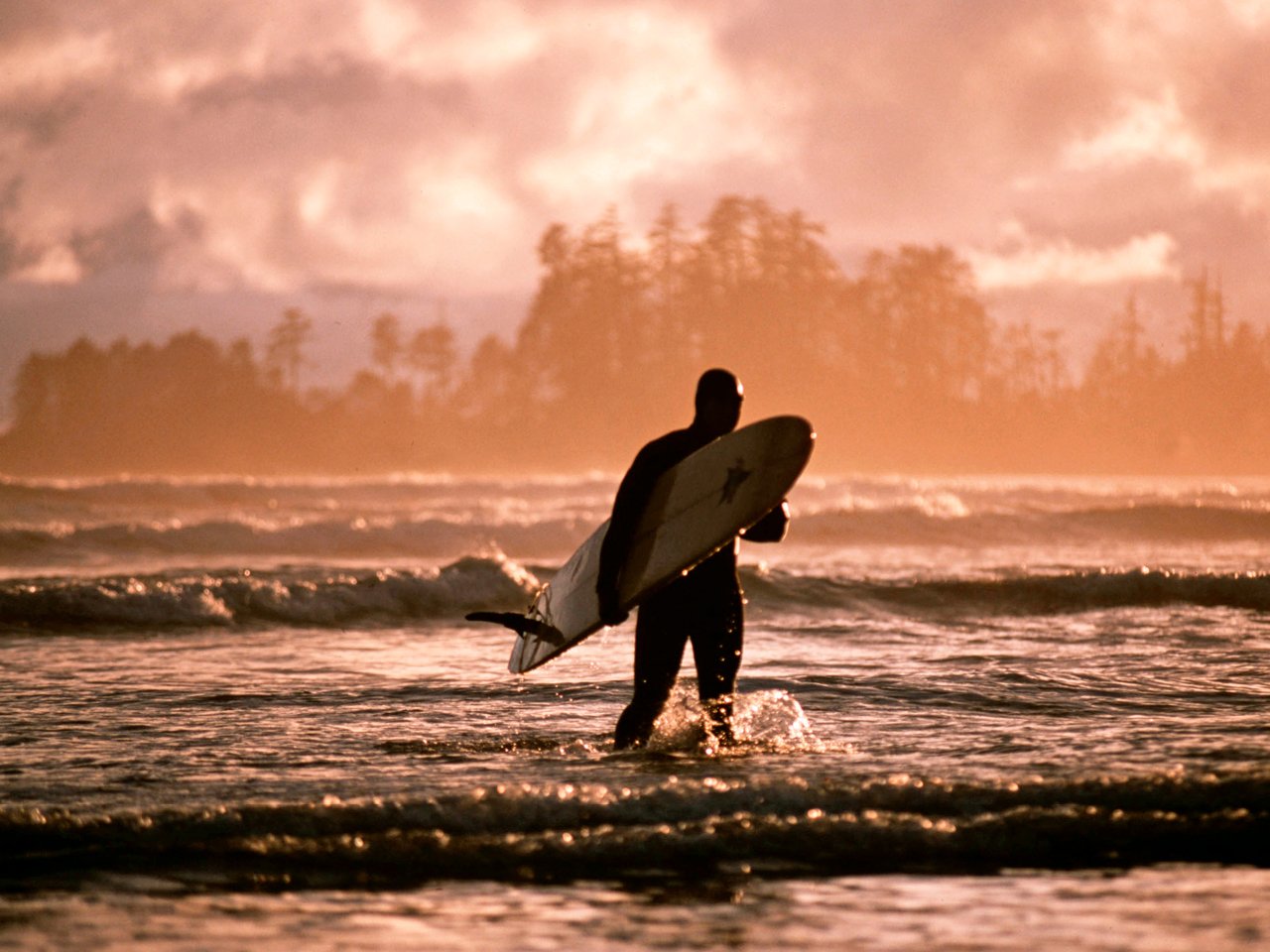 Full body suited surfer, board in hand, walks through low-tide