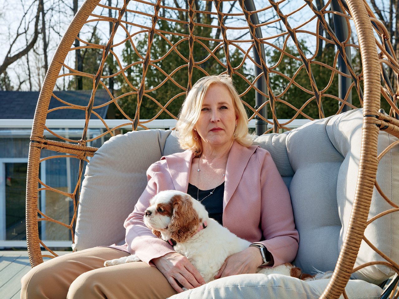 LIsa Raitt in a black shirt and pink blazer, sitting on a patio chair with her king Cavalier spaniel in her lap