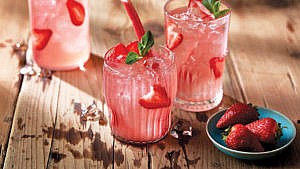Three glasses of strawberry rhubarb sangria, a pink drink with floating strawberries, on a wood table beside a plate of cut strawberries