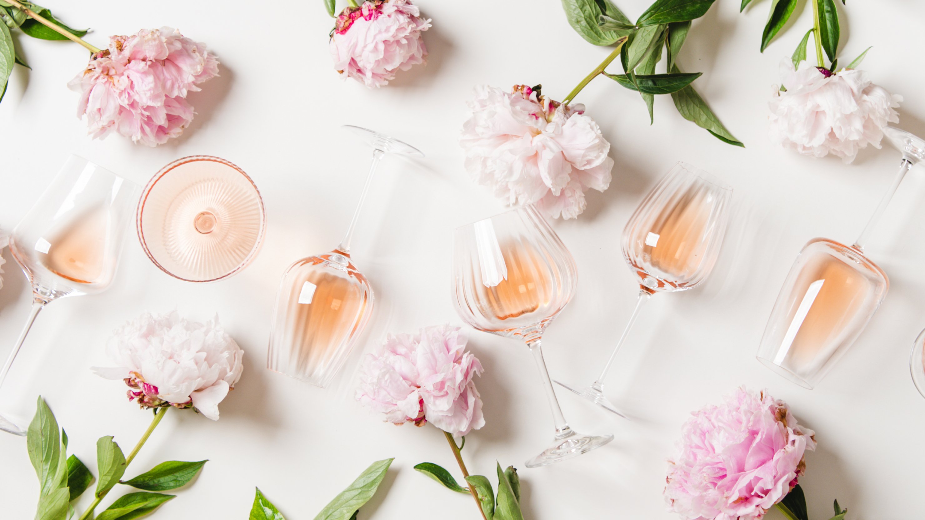 Flat-lay of rose wine in glasses and summer peony flowers over white background, top view.
