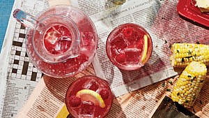 A pitcher of ice-filled raspberry cordial and two glasses, garnished with lemons, on a newspaper backdrop