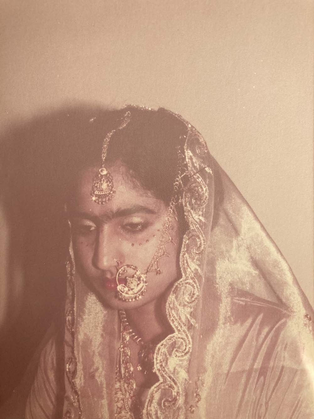 An old photo of the author's mother at a wedding, wearing red and gold jewellery, including nath (a nose ring and chain)
