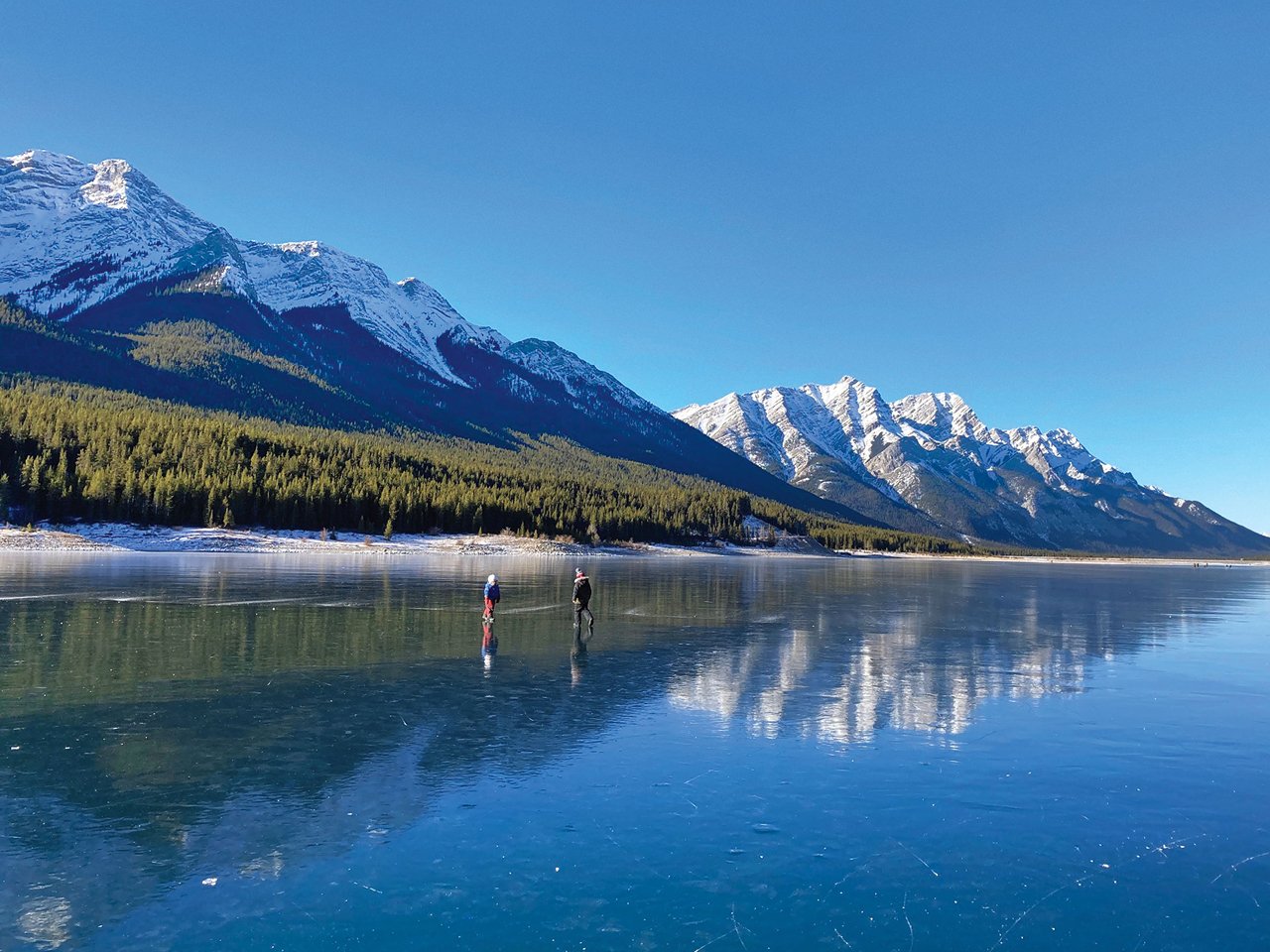 Two people skate on a lake with the Rockies and blue sky in the background