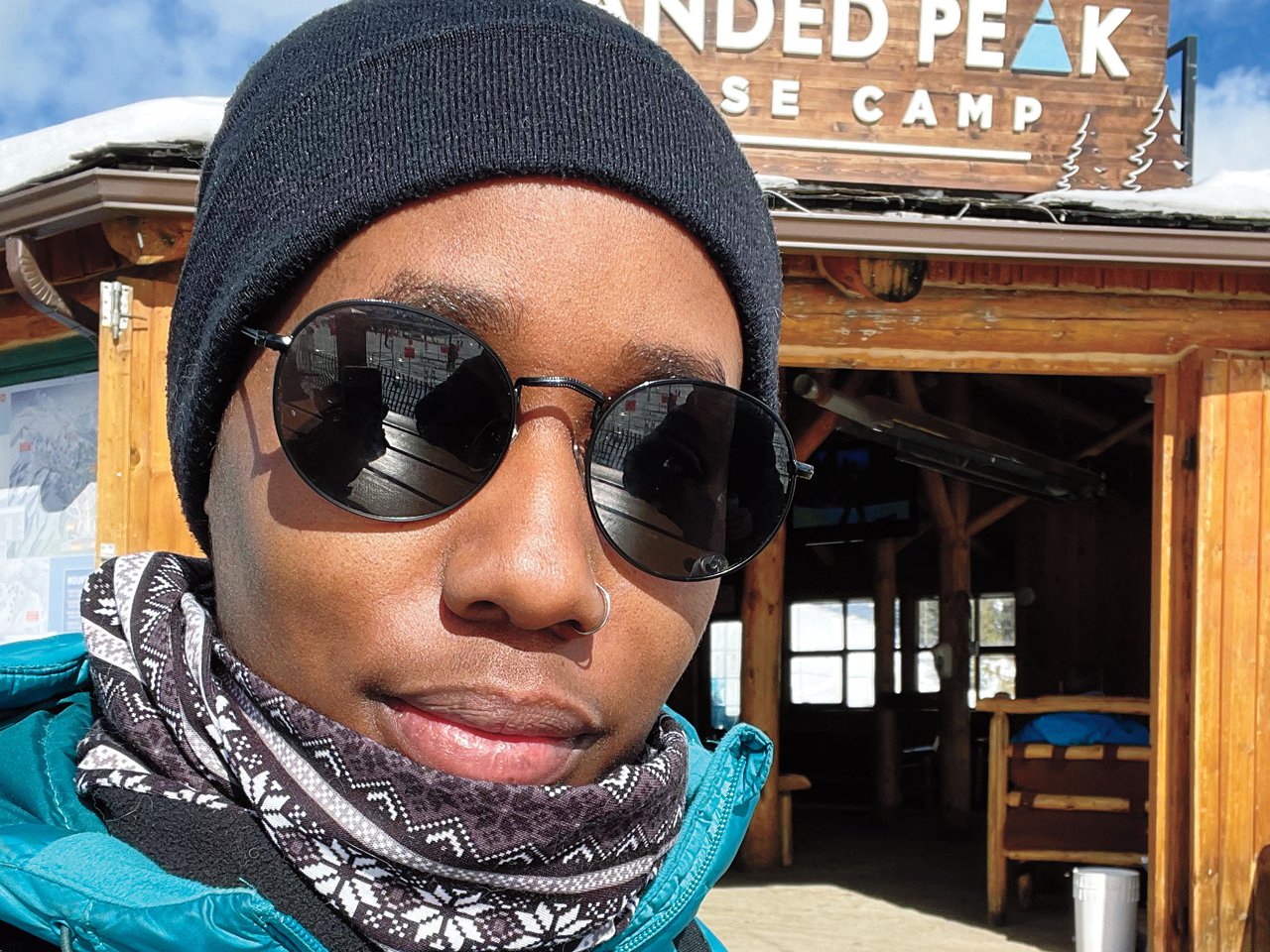 A photo of a Black woman in a black tuque and turquoise ski jacket in front of a ski chalet