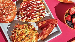 An okonomiyaki breakfast sandwich on a plate with bacon, with a side bowl of berries and a glass of orange juice