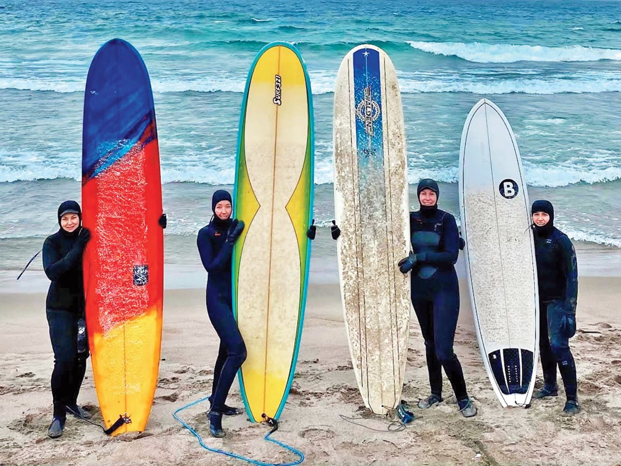 Four women in wetsuits holding surfboards standing on a beach in front of a lake