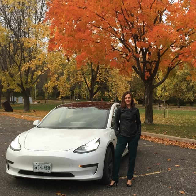 Laura Mason stands beside her white Tesla Model 3 Standard Range Plus in front of a fall tree backdrop