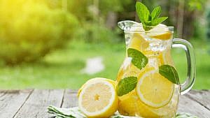 A pitcher of lemonade filled with ice on a wood table next to a sliced lemon against a backdrop of shrubs and grass