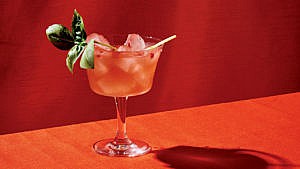 Gin con Tomate cocktail sits in front of red backdrop, there is a basil garnish popping out of the top of the glass