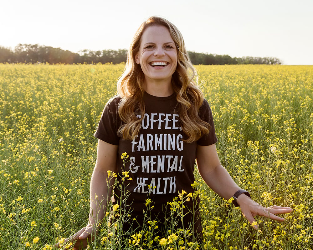 Lesley Kelly stands in a field of flowers, her shirt says 'Coffee, Farming and Mental Health'