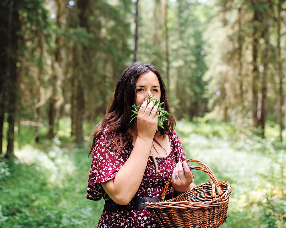 Tiffany Traverse stands in a forest, holding a wicker basket in her left hand and a handful of plants up to her nose in her right hand