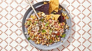 A bowl of Mexican street corn salsa with a spoon resting in it and tortilla chips at the side on a patterned table