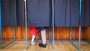 A photo of a curtained voting booth, with the feet of a small child in a red coat and a parent sticking out under the curtain