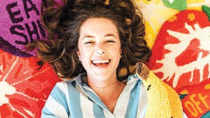 A woman smiling against a background of colourful rugs to illustrate an article on bold rug hooking in Nova Scotia.