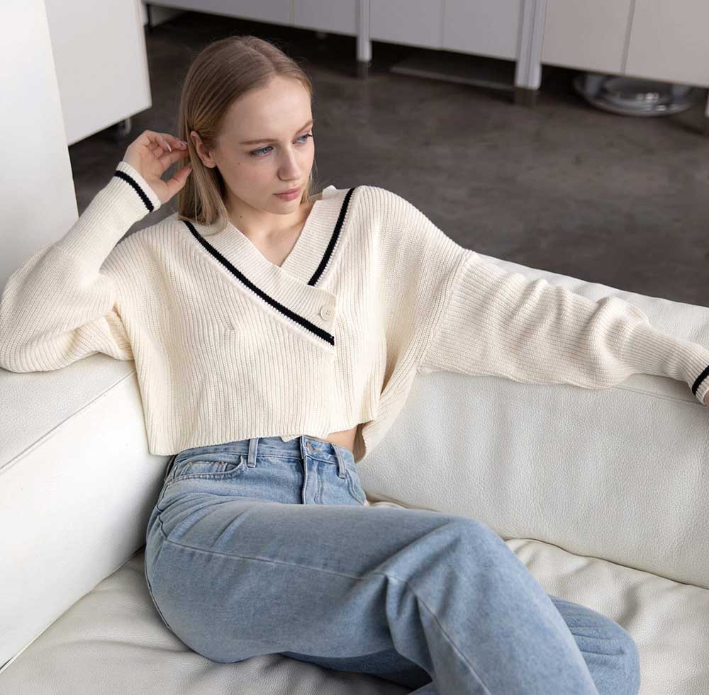 A model sitting on a couch wearing a white cropped varsity-style cardigan with black collar detailing from Oak and Fort.
