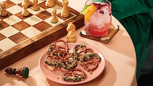 A plate of chocolate covered pretzels on a table next to a chess board and a drink