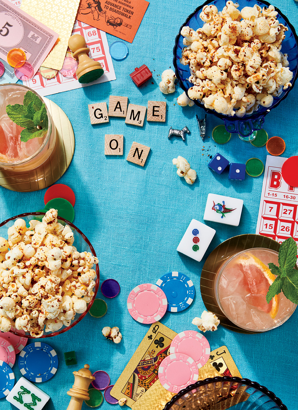 A furikake popcorn bowl, a grapefruit and sake cocktail, next to an array of game supplies (chess, cards, chips) with Scrabble letters spelling out "Game on" on a blue tablecloth