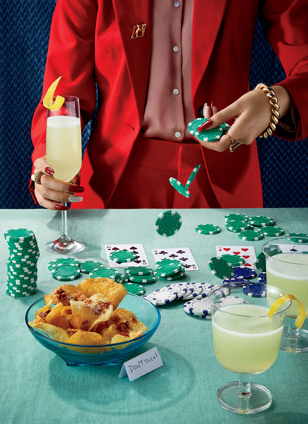 A woman in a red suit with red nails holds a limoncello prosecco cocktail with a lemon garnish, she is dropping game chips onto a table laden with cards and parmesan and parma ham chips, it has a green tablecloth