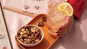 A bowl of warm sweet and savour nuts on an orange tray, beside a hand holding a whisky and tonic in a tall glass with a lemon garnish
