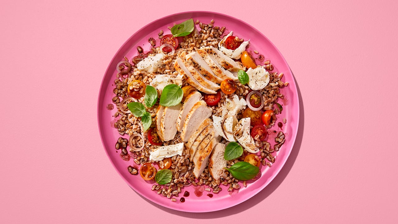 A pink bowl of farro salad with chicken and caprese on a pink table.