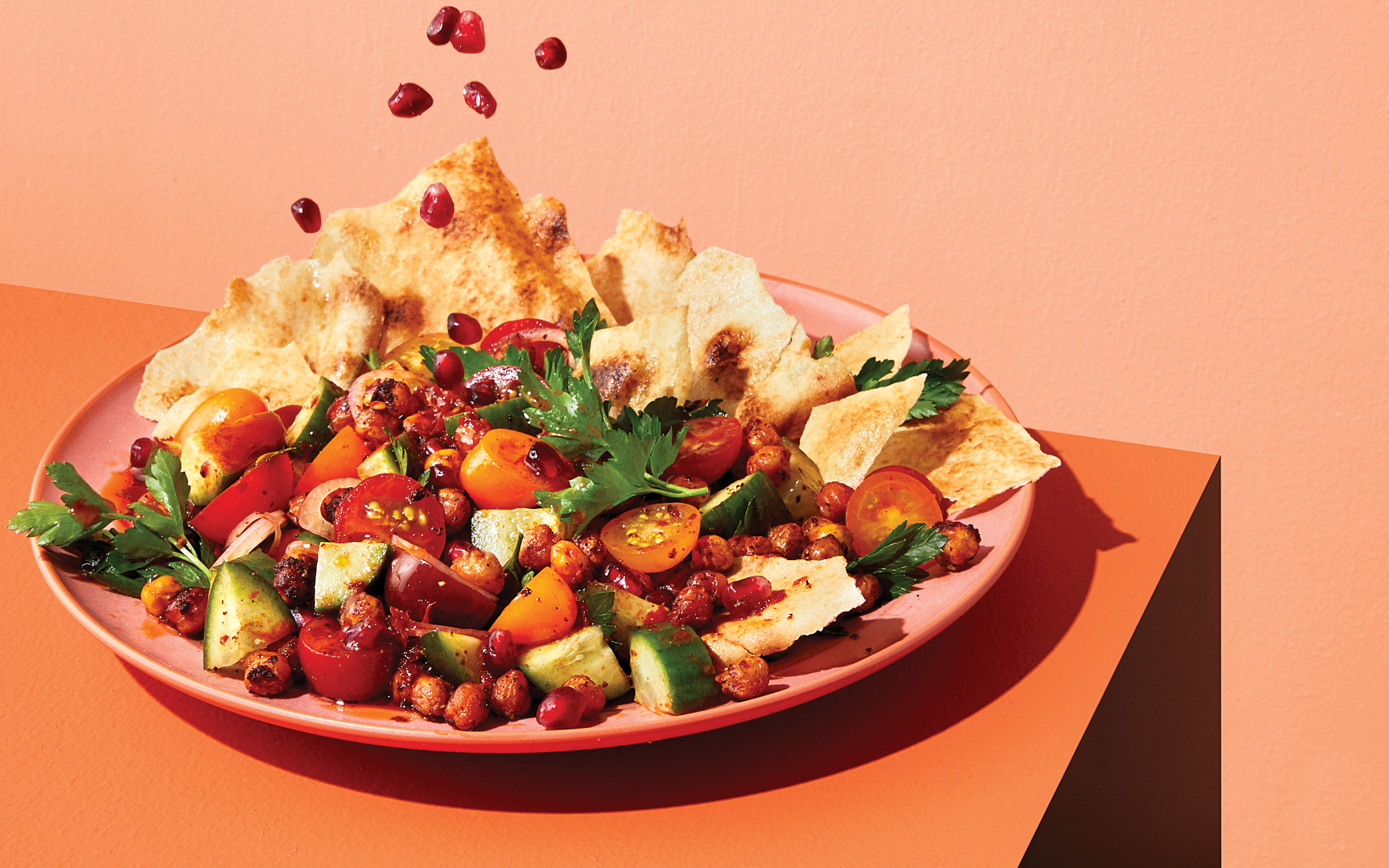 A salad with cucumbers, tomatoes, pita chips and chickpeas on a orange table against an orange background.