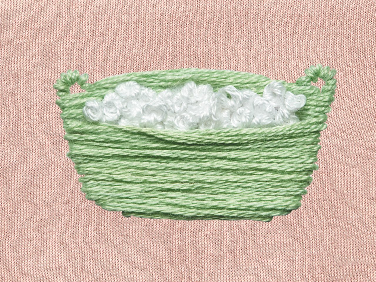 An embroidered green laundry basket with white suds on a pink cloth background