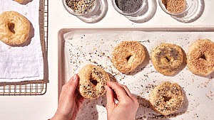 A photo of a person's hands covering homemade bagel in a mixture of sesame and poppy seeds